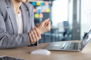 A woman is pictured below the shoulder holding her wrist while sitting at a desk with a keyboard and mouse.